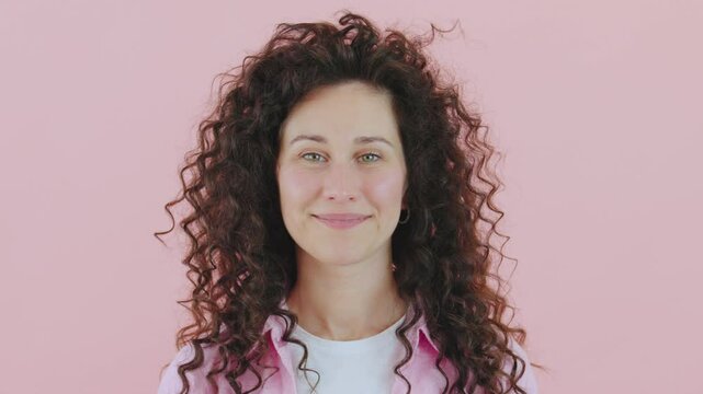 Woman with curly hair stands in front of a pink background, eyes closed,opening them and smiling, practicing mindfulness and meditative breathing, enjoying a moment of calm and tranquility