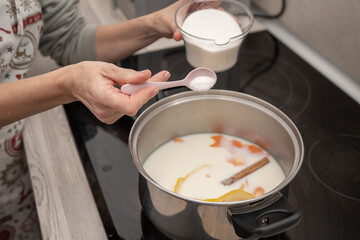 Woman adding sugar to a pot of milk with citrus peels and cinnamon on the stove