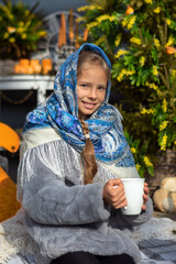 A beautiful Russian girl in a fur coat and headscarf sits on the porch of a wooden house on a sunny spring day. Family values, traditions and home comfort.Tea drinking outside the house