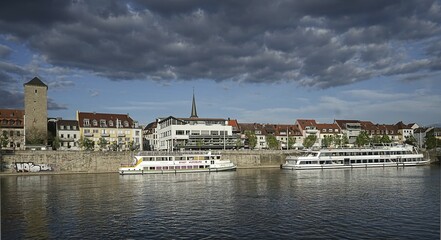 Two boats moored on a riverside promenade with historic buildings and a striking tower under a cloudy sky Würzburg am Main Germany