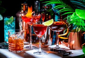 Summer alcoholic cocktail party with ready-made cocktails, bar tools and bottles of spirits on marble table with tropical plants. Green background, hard light and shadows. Shallow DOF