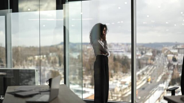 Confident young businesswoman talking on the phone and looking out the window of modern office building. Successful female executive having phone call in office.