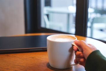 A hand holding a cup of coffee with laptop on the table