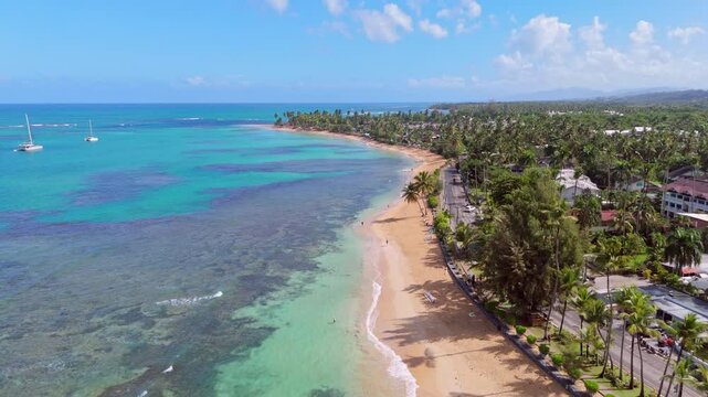 Playa Punta Popy Sandy beach during sunny day with coral reef water. Aerial top down flyover coastline. Driving cars on coastal street with tropical palm trees. Wide shot.
