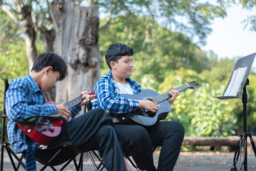 Fototapeta premium Asian teenaged boy spending his free times with his friend on weekend by practising and playing acoustic guitar or ukulele in the park together, recreational activity of teenagers concept.