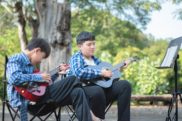 Fototapeta premium Asian teenaged boy spending his free times with his friend on weekend by practising and playing acoustic guitar or ukulele in the park together, recreational activity of teenagers concept.