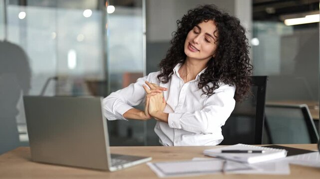 Beautiful businesswoman with curly hair stretching her arms and back at the office desk. She takes a deep breath and smiles.