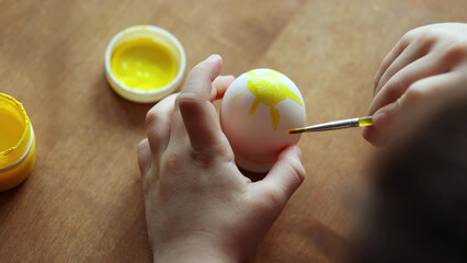child paints yellow sun on white egg with paint and brush for Easter at home at table. close-up, top view