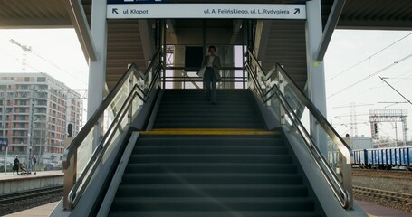 Obraz premium A woman in formal clothes and headphones, with a cup of coffee in her hand, descends the stairs to the platform of the railway station