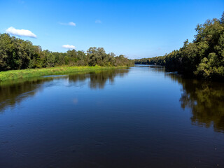 A calm river with trees on both sides. The water is clear and the sky is blue. The scene is peaceful and serene