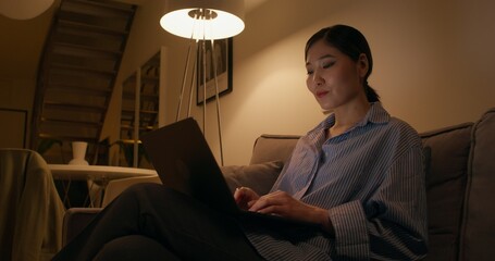 A woman puts on headphones and typing on a laptop sitting on the couch in an apartment, dimmed...