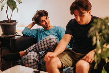 Two young men are sitting on a couch, engaged in a serious discussion. The indoor setting features...