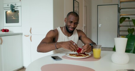 A man in pajamas squeezes jam out of a package onto pancakes while having breakfast, sitting at the table at home. Daylight