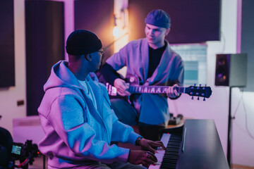 Two musicians, one playing the piano and the other guitar, work together in a studio. The dim...
