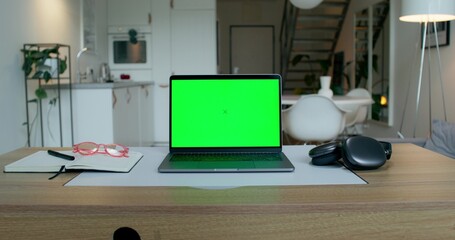 A laptop with a green screen is on the desktop at home. An open notebook and headphones next to it