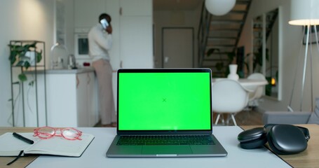 A laptop with a green screen is on the desktop at home. A man is talking on a mobile phone standing in the kitchen in the background