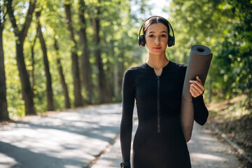 Model is with yoga mat. Young sportive skinny woman is in black fitness clothes outdoors