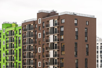 Modern apartment buildings with green and brown facades, large windows, and balconies with metal railings, standing against a cloudy sky, urban living