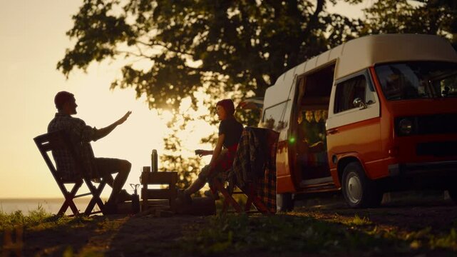 Calmness and relaxation in nature, campers traveling across country by RV. Silhouette view of two tourists sitting at small table on coast of lake or sea in sunset, slow motion shot, happy people