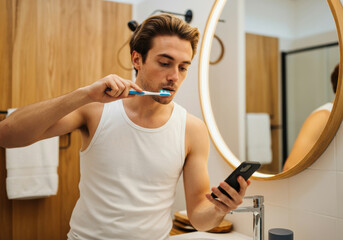 Young man brushes teeth while using smartphone in modern bathroom during morning routine