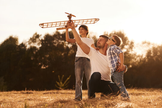 Family of father, son and daughter are playing with wooden toy plane on the field - Powered by Adobe