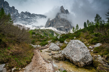 Rocky hiking trail near Lago Sorapis in the Dolomites (Dolomiti, Dolomiten), Italy. The path runs alongside a small mountain stream, with massive boulders and peaks in the background