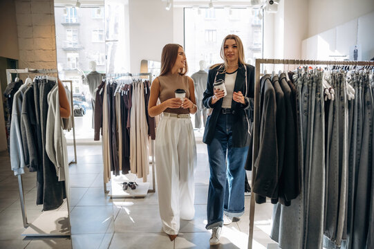 Cups of drinks in hands. Two female friends are in the retail store, choosing clothes, shopping