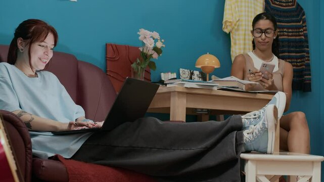 Two cheerful female students searching information on internet using laptop and smartphone while completing homework assignment in dorm room