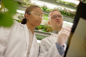 Two scientists wearing white lab coats and safety goggles examining data on large tablet device...