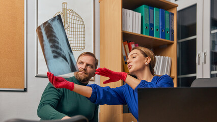 Woman, doctor in blue medical coat sitting at table and looking at x-ray with bearded man, patient....