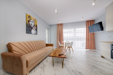 Salon w nowoczesnym apartamencie z beżowym  aneksem kuchennym z rudą jasno brązową pluszową sofą
