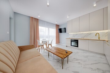 Salon w nowoczesnym apartamencie z beżowym  aneksem kuchennym z rudą jasno brązową pluszową sofą