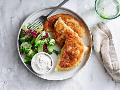 A plate features homemade chebureki, golden brown and crispy, alongside a fresh green salad and creamy dipping sauce. This appetizing meal captures the joy of homemade cooking