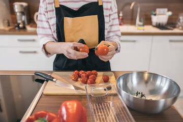 Girl wearing an apron holding cherry tomatoes over a wooden cutting board in a modern kitchen, preparing a healthy meal