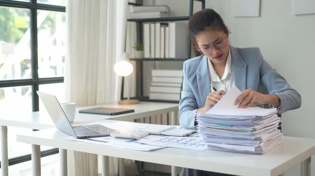 A woman is sitting at a desk with a pile of papers in front of her. She is wearing a blue jacket and glasses. The scene suggests that she is working on a project or dealing with paperwork