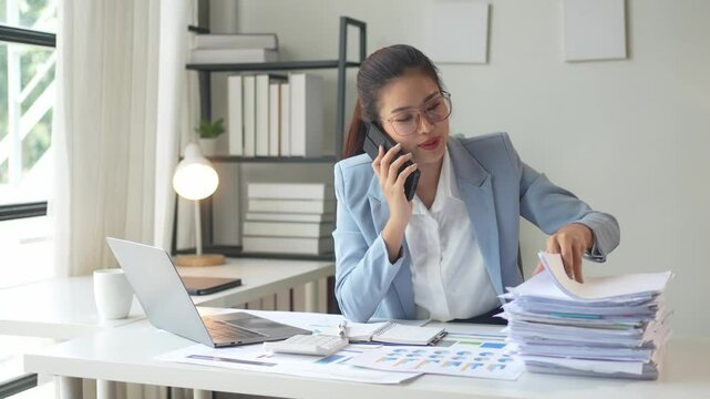 A woman is talking on her cell phone while sitting at a desk with a pile of papers in front of her