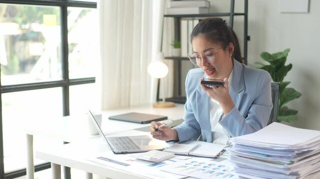 A woman in a business suit is talking on her cell phone while sitting at a desk with a laptop and a stack of papers