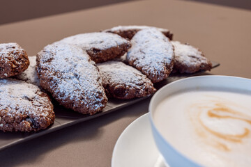 Freshly baked Pan dei Morti cookies with powdered sugar displayed on a textured plate, paired with creamy cappuccino in a warm, inviting setting