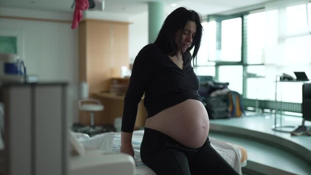 Struggling pregnant woman seated by bedside of hospital bed having contractions during stages of labor, 30s expecting lady suffering from pain during childbirth