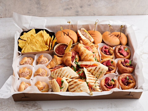 A variety of gourmet snacks including sandwiches, pastries, and chips served in a decorative box, perfect for gatherings or parties. Enjoy the colorful presentation and delectable flavors