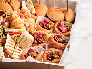 A variety of delectable savory treats is displayed in a well-organized box, featuring sandwiches, pastries, and meats garnished with herbs, perfect for any occasion