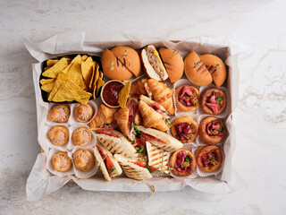 A variety of savory snacks fills a rectangular tray, featuring sliders, pastries, chips, and dips, perfect for a casual gathering or celebration