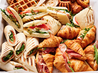 A variety of sandwiches, croissants, and pastries are beautifully arranged for a brunch. Fresh ingredients include greens and rich spreads, appealing to all tastes