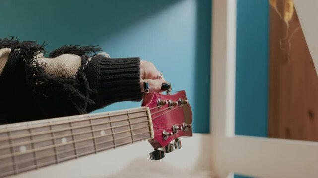 Close-up view of unrecognisable girl tuning acoustic guitar strings by adjusting pegs in dormitory room