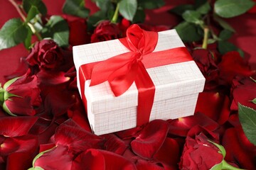 Beautiful gift box among rose flowers and petals on red background, closeup. Romantic present