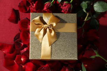 Beautiful gift box among rose flowers on red background, flat lay. Romantic present
