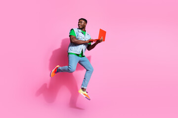 Young, stylish man with a vibrant personality jumps against a pink background, using a laptop.