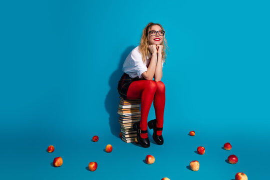 Blond woman in a school uniform sitting on books with apples around on a vibrant blue background