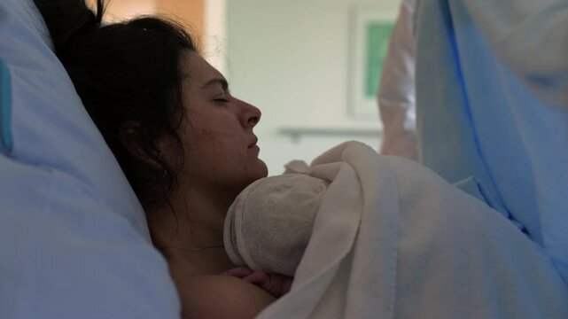 New mother struggling with pain after childbirth laid in bed being taken care by medical team, covering her blankets while she holds her newborn baby close to her chest