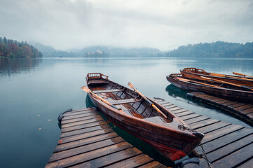 Wet boats tied to a wooden pier in a rainy day on Lake Bled against the backdrop of the beautiful church on the island in the middle of the lake, Julian Alps, Slovenia